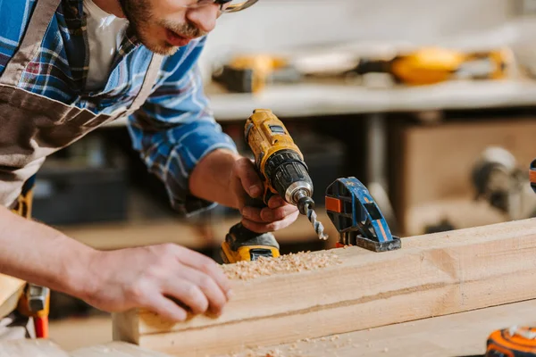 Cropped view of bearded carpenter in apron holding hammer drill near wooden planks — Stock Photo