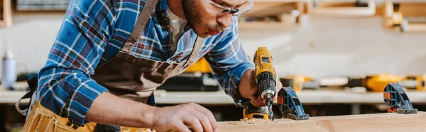 Panoramic shot of carpenter in safety glasses holding hammer drill and blowing on sawdust near wooden plank — Stock Photo