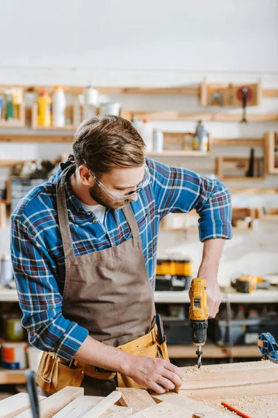 Selective focus of woodworker in safety glasses and apron holding hammer drill near wooden planks — Stock Photo