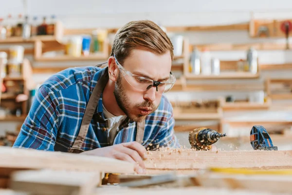 Selective focus of carpenter in goggles holding hammer drill and blowing on sawdust near wooden plank — Stock Photo