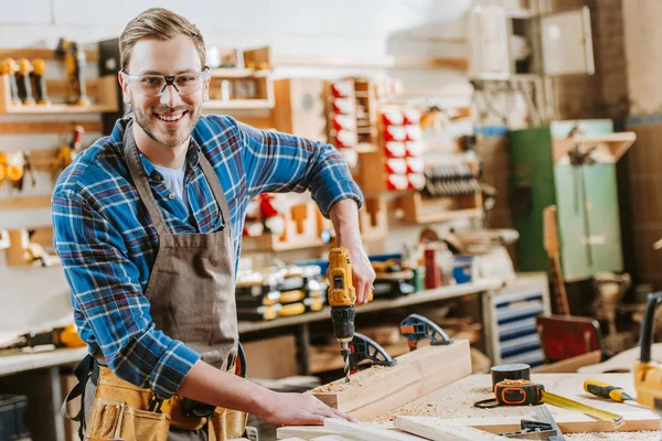 Happy woodworker in goggles and apron holding hammer drill near wooden planks — Stock Photo
