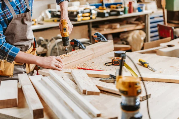 Vista recortada del carpintero en delantal sosteniendo taladro de martillo cerca de tablones de madera en el taller - foto de stock