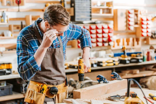 Selective focus of woodworker in apron touching goggles and holding hammer drill near wooden planks — Stock Photo