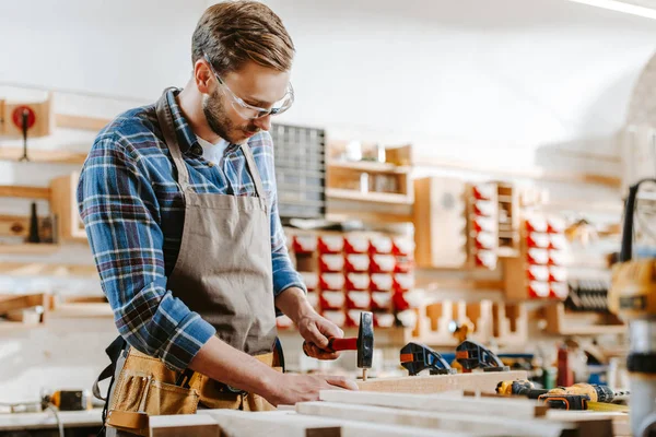 Selective focus of carpenter in goggles holding hammer near wooden dowel in workshop — Stock Photo