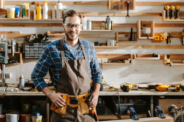 Cheerful carpenter in goggles standing and touching tool belt in workshop — Stock Photo