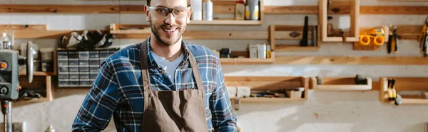 Panoramic shot of cheerful carpenter in apron looking at camera in workshop — Stock Photo