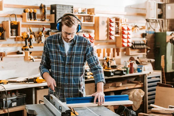 Handsome woodworker in protective headphones holding plank near circular saw — Stock Photo