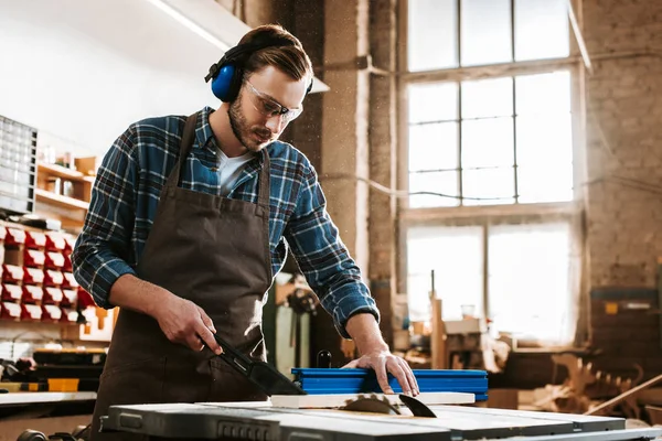 Beau menuisier en casque protecteur et tablier tenant la planche près de la scie circulaire — Photo de stock