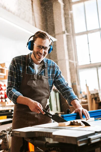 Cheerful woodworker in protective headphones and apron holding plank near circular saw — Stock Photo