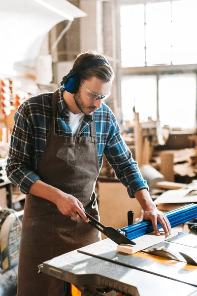 Bearded woodworker in protective headphones and apron holding plank near circular saw — Stock Photo