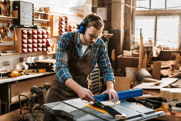 Handsome woodworker in protective headphones and apron holding plank near circular saw — Stock Photo