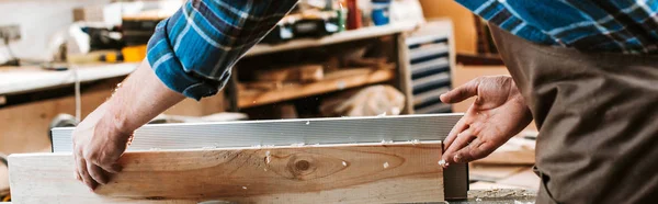 Panoramic shot of woodworker holding plank near circular saw in carpentry shop — Stock Photo