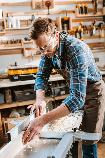 Woodworker in apron and goggles holding plank near circular saw — Stock Photo