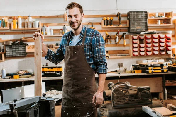 Fröhlicher Tischler in Schürze steht und hält Holzplanke in Werkstatt — Stockfoto