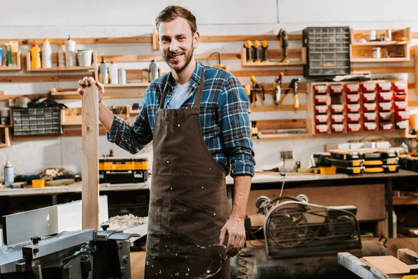 Charpentier joyeux dans le tablier debout et tenant la planche en bois dans l'atelier — Photo de stock