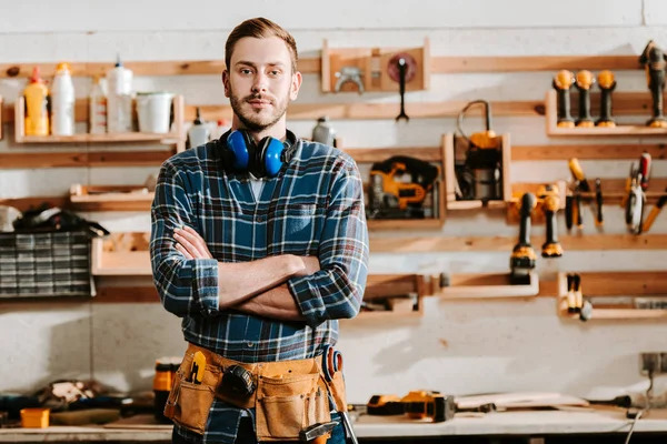 Handsome carpenter in apron standing with crossed arms in workshop — Stock Photo