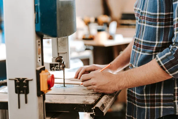 Cropped view of carpenter holding pencil near electric woodworking in workshop — Stock Photo