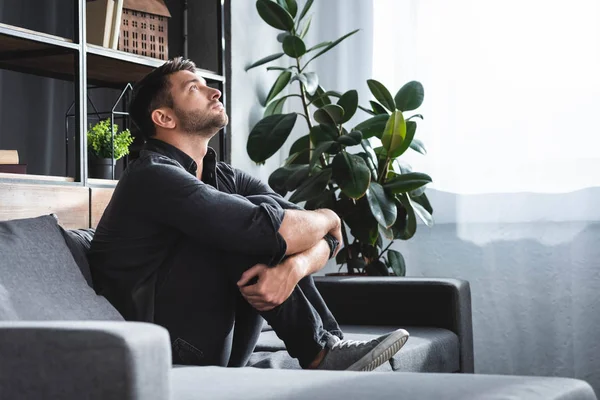 Vista lateral del hombre guapo con ataque de pánico sentado en el sofá y abrazando las piernas en el apartamento - foto de stock