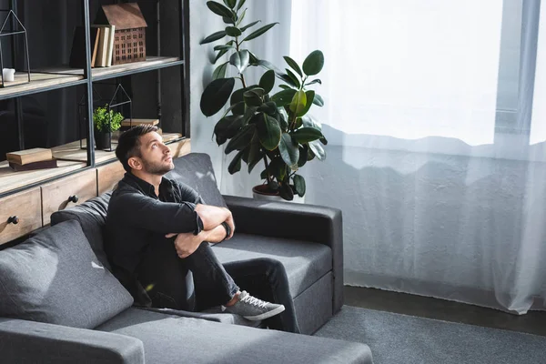 Blick aus der Vogelperspektive auf einen gutaussehenden Mann mit Panikattacke, der auf dem Sofa sitzt und die Beine in der Wohnung umarmt — Stockfoto