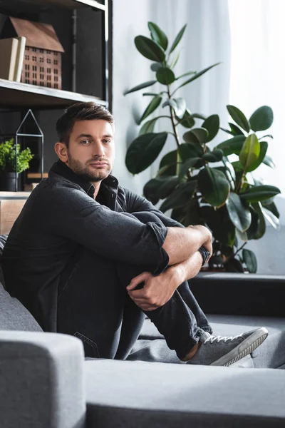 Schöner Mann mit Panikattacke sitzt auf Sofa und umarmt Beine in Wohnung — Stockfoto