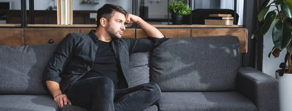 Tiro panorâmico de homem bonito e pensativo sentado no sofá no apartamento — Fotografia de Stock