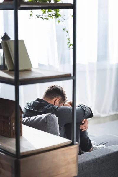 Vista trasera del hombre con ataque de pánico llorando y abrazando las piernas en el apartamento - foto de stock