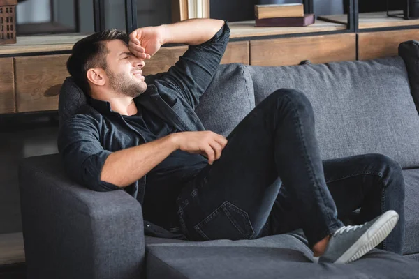 Hombre guapo con dolor de cabeza sentado en el sofá y tocando la cabeza en el apartamento - foto de stock