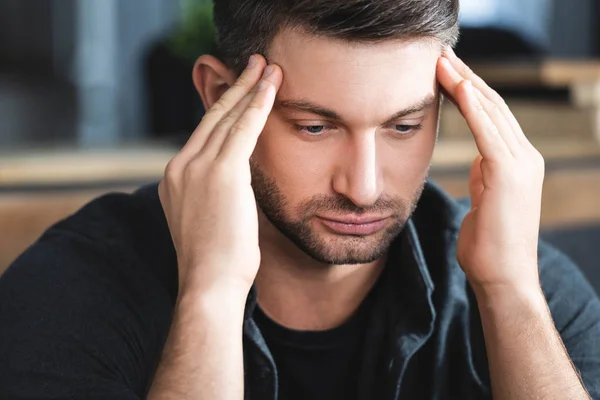 Hombre guapo con dolor de cabeza tocando la cabeza y mirando hacia abajo en el apartamento - foto de stock