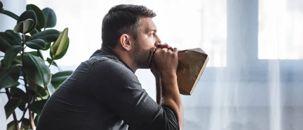 Panoramic shot of handsome man with panic attack breathing in paper bag in apartment — Stock Photo