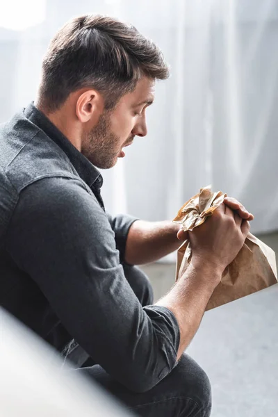 Selective focus of handsome man with panic attack holding paper bag in apartment — Stock Photo