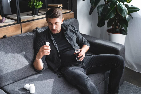 Vue grand angle de bel homme avec attaque de panique tenant des pilules et un verre d'eau dans l'appartement — Photo de stock