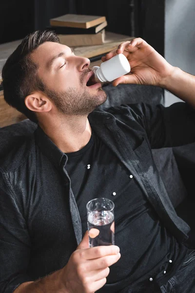 Vista de ángulo alto del hombre guapo con ataque de pánico tomando pastillas y sosteniendo un vaso de agua en el apartamento - foto de stock