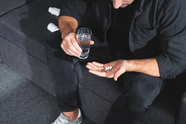 Vista ritagliata dell'uomo con attacco di panico in possesso di pillole e bicchiere d'acqua in appartamento — Foto stock
