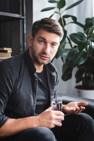 Schöner Mann mit Panikattacke hält Tabletten und Glas Wasser in Wohnung — Stockfoto