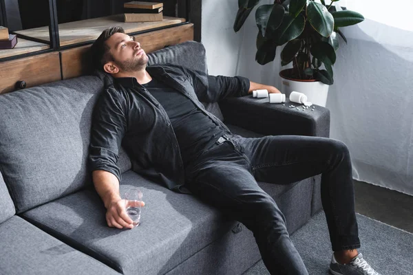 Vista de ángulo alto del hombre guapo con ataque de pánico sosteniendo un vaso de agua y sentado en el sofá en el apartamento - foto de stock