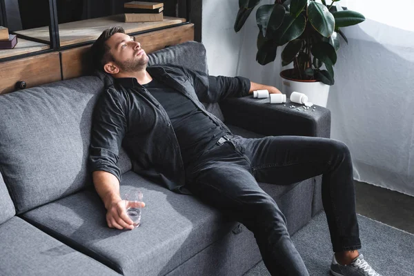 High angle view of handsome man with panic attack holding glass of water and sitting on sofa in apartment — Stock Photo