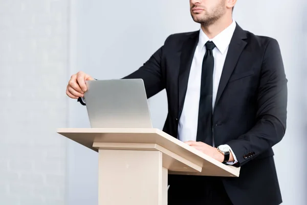 Vista recortada de hombre de negocios en traje de pie en el tribuno podio y portátil de apertura durante la conferencia aislado en blanco - foto de stock