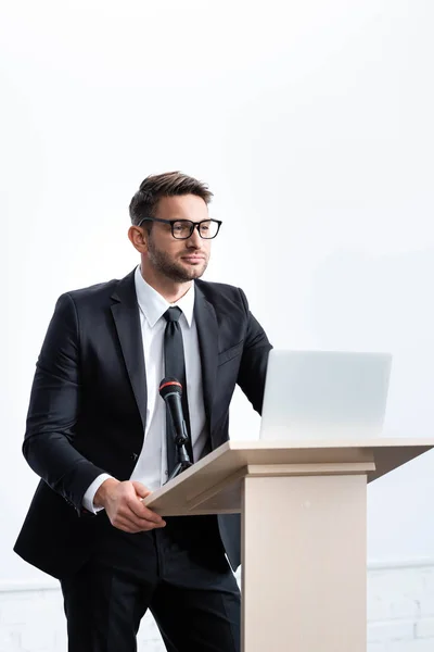 Бизнесмен в костюме стоит на трибуне и смотрит в сторону во время конференции изолированы на белом — стоковое фото