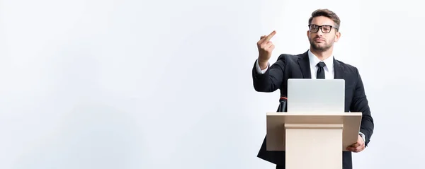 Panoramic shot of businessman in suit standing at podium tribune and showing middle finger during conference isolated on white — Stock Photo