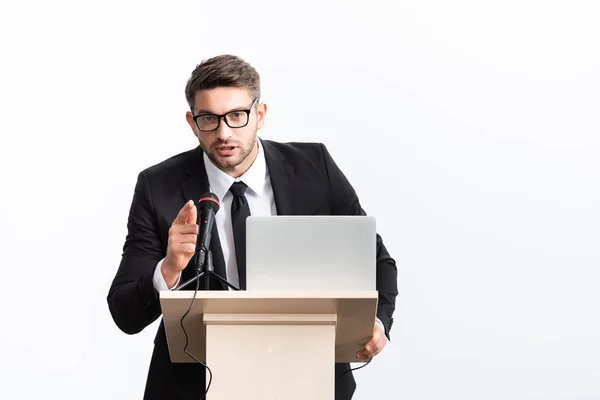 Businessman in suit standing at podium tribune and pointing with finger during conference isolated on white — Stock Photo