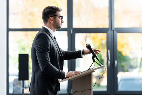 Vue de côté de l'homme d'affaires en costume debout sur le podium tribune et regardant loin pendant la conférence — Photo de stock