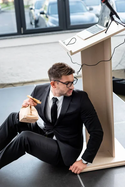 Ängstlicher Geschäftsmann im Anzug hält Papiertüte in der Hand und versteckt sich während der Konferenz hinter der Tribüne — Stockfoto