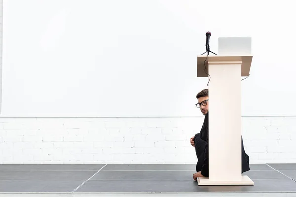 Наляканий бізнесмен в окулярах ховається за подіумом трибуни під час конференції ізольовані на білому — стокове фото