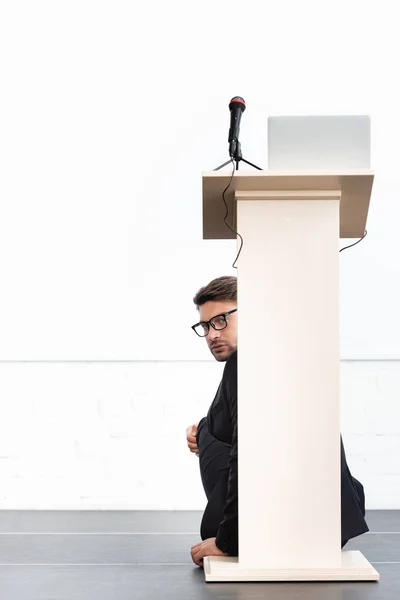 Scared businessman in glasses hiding behind podium tribune during conference isolated on white — Stock Photo