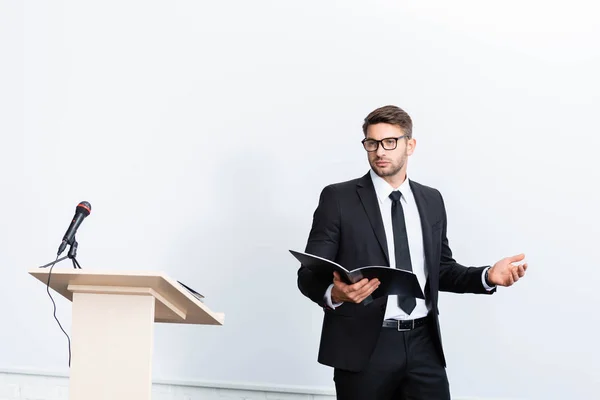 Hombre de negocios en traje sosteniendo la carpeta y mirando hacia otro lado durante la conferencia sobre fondo blanco - foto de stock