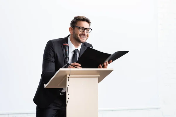 Улыбающийся бизнесмен в костюме, стоящий на трибуне и держащий папку во время конференции изолированы на белом — стоковое фото