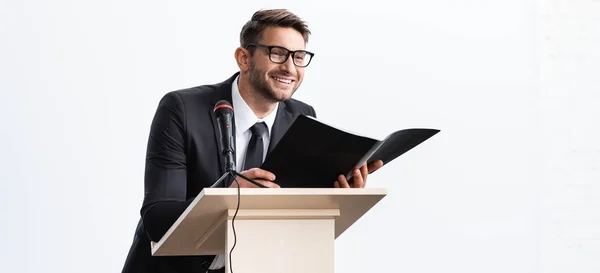 Plano panorámico de sonriente hombre de negocios en traje de pie en el tribuno podio y la celebración de la carpeta durante la conferencia aislado en blanco - foto de stock