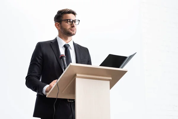 Businessman in suit standing at podium tribune and looking away during conference isolated on white — Stock Photo