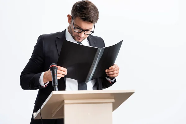 Businessman in suit standing at podium tribune and holding folder during conference isolated on white — Stock Photo