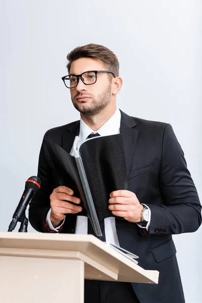 Businessman in suit standing at podium tribune and holding folder during conference isolated on white — Stock Photo